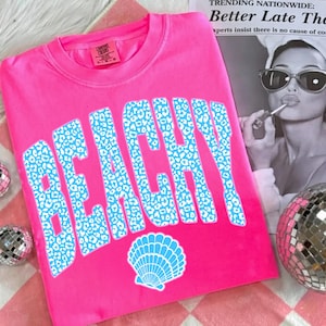 May include: A bright pink t-shirt with the word "BEACHY" in large, light blue leopard print letters. A light blue seashell graphic is below the word. The shirt is a casual style.