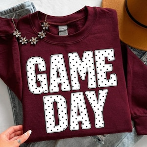 May include: A maroon sweatshirt with the words "GAME DAY" in large white letters with black polka dots. A silver necklace with flower-shaped accents is draped over the sweatshirt.