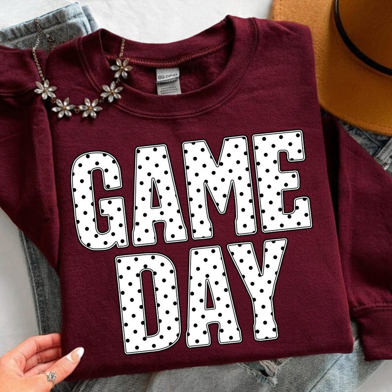 May include: A maroon sweatshirt with the words "GAME DAY" in large white letters with black polka dots. A silver necklace with flower-shaped accents is draped over the sweatshirt.
