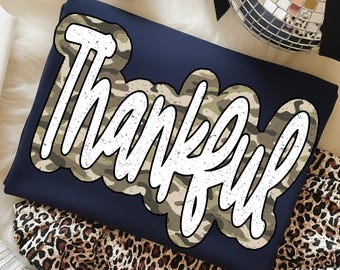 Könnte beinhalten: Marineblaues Sweatshirt mit dem weißen Wort "Thankful", umrandet von einem Camouflagemuster. Das gefaltete Kleidungsstück liegt auf einer weißen Oberfläche, mit Leopardenmuster-Stoff.