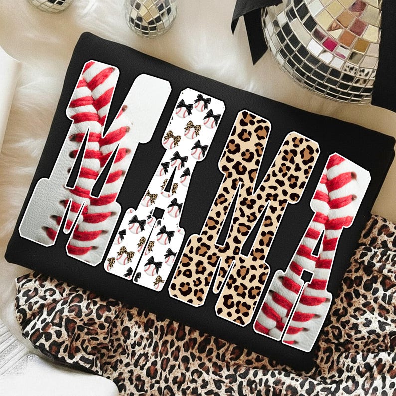 May include: Black sweatshirt with the word "MAMA" in a unique design. The letters are filled with baseball stitching, baseballs with bows, and leopard print. The sweatshirt is on a leopard print fabric.