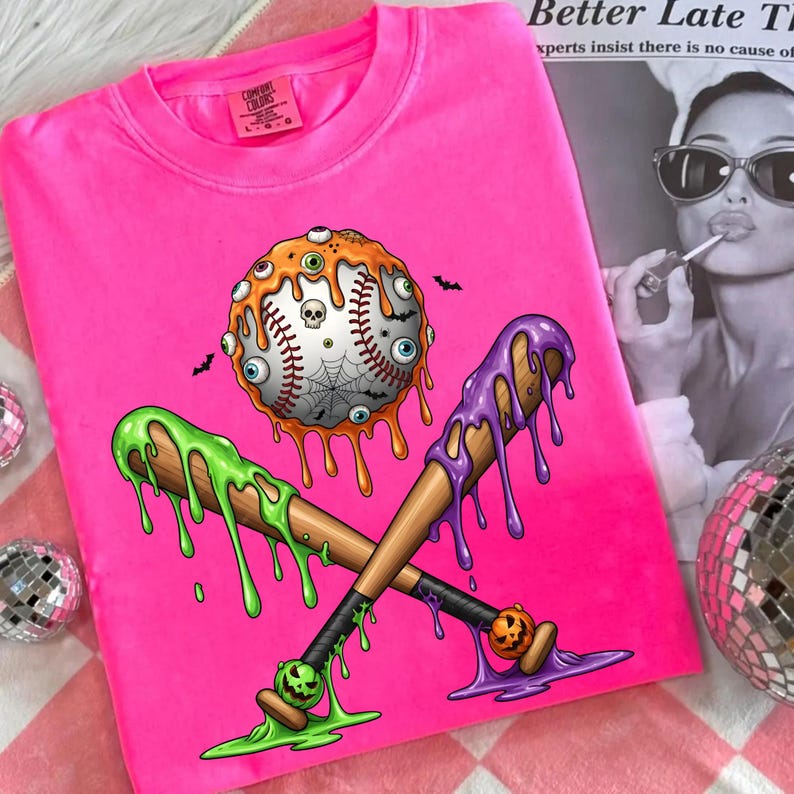 K&ouml;nnte beinhalten: Leuchtend pinkes T-Shirt mit Halloween-Motiv. Das Design zeigt einen Baseball, der mit Aug&auml;pfeln, einem Spinnennetz und einem Totenkopf bedeckt ist, mit gekreuzten Baseballschl&auml;gern, die mit gr&uuml;nem und lila Schleim tropfen. Die Schl&auml;ger haben K&uuml;rbislaternen.