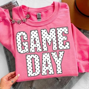 May include: A pink sweatshirt with the words "GAME DAY" in large, white, polka-dotted letters. A floral necklace is draped across the top. The sweatshirt is folded on top of a pair of gray jeans.