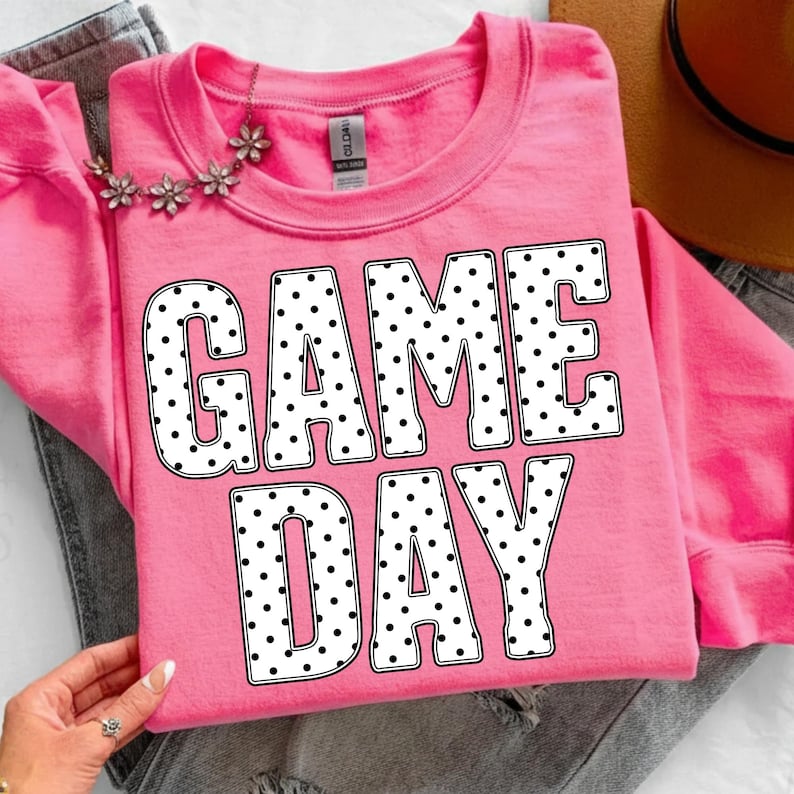 May include: A pink sweatshirt with the words "GAME DAY" in large, white, polka-dotted letters. A floral necklace is draped across the top. The sweatshirt is folded on top of a pair of gray jeans.