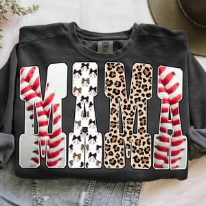 May include: A dark gray sweatshirt with the word "MAMA" in large, bold letters. The letters are decorated with baseball stitching, bows, and leopard print. The sweatshirt is a Comfort Colors brand.