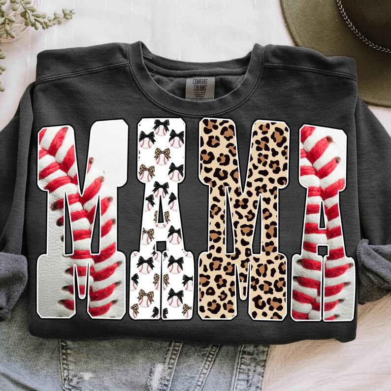 May include: A dark gray sweatshirt with the word "MAMA" in large, bold letters. The letters are decorated with baseball stitching, bows, and leopard print. The sweatshirt is a Comfort Colors brand.