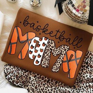 May include: A brown sweatshirt with the words "basketball mama" in black script. The word "mama" is designed with basketballs, leopard print, and basketballs with bows. The sweatshirt is folded on a leopard print fabric.