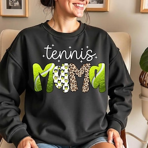 K&ouml;nnte beinhalten: Schwarzes Sweatshirt mit dem Schriftzug "tennis mama" in wei&szlig;er Schrift. Das Wort "mama" ist mit einem Leopardenmuster und Tennisball-Grafiken gestaltet. Das Sweatshirt hat einen Rundhalsausschnitt und lange &Auml;rmel.