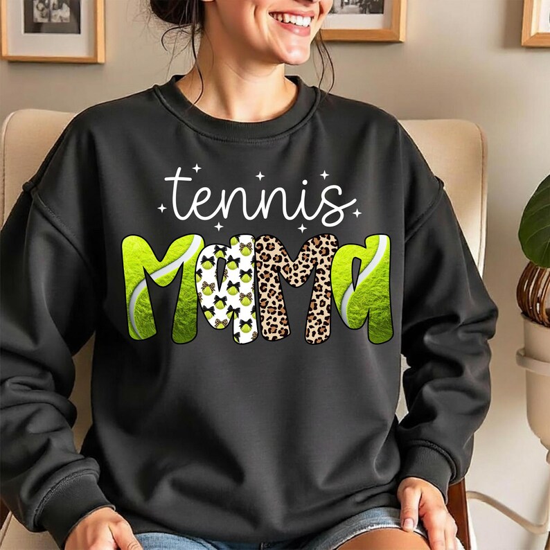 K&ouml;nnte beinhalten: Schwarzes Sweatshirt mit dem Schriftzug "tennis mama" in wei&szlig;er Schrift. Das Wort "mama" ist mit einem Leopardenmuster und Tennisball-Grafiken gestaltet. Das Sweatshirt hat einen Rundhalsausschnitt und lange &Auml;rmel.