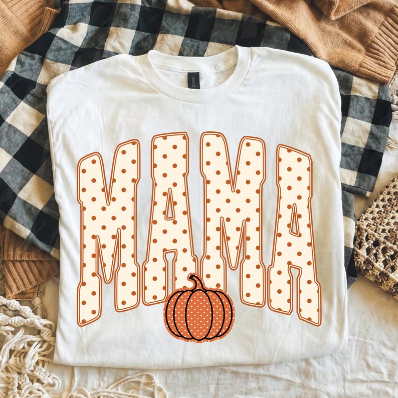 May include: A white short-sleeved t-shirt featuring the word MAMA in large, block-style letters with a cream background and brown polka dots. Below the text is a graphic of a pumpkin with a matching cream and brown polka dot pattern.