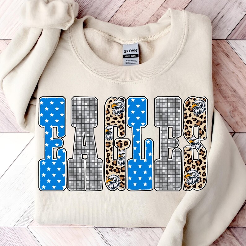 May include: Cream-colored sweatshirt with the word "EAGLES" in a bold, block letter design. The letters feature a mix of blue star patterns, leopard print, and a mosaic-like pattern. Eagle head graphics are incorporated.