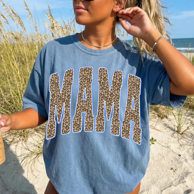 May include: A blue short-sleeved t-shirt featuring the word MAMA printed in a large, bold, arched font. The letters have a brown and black leopard print pattern with a thin white outline, creating a high-contrast graphic on the solid blue fabric.