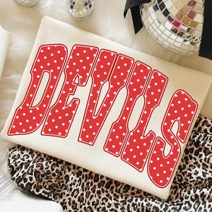 Puede incluir: Una sudadera de color crema con la palabra DEVILS en letras rojas grandes y llamativas con un patrón de lunares blancos. El texto tiene un borde rojo grueso y está colocado en el centro de la parte delantera de la prenda, que está doblada.