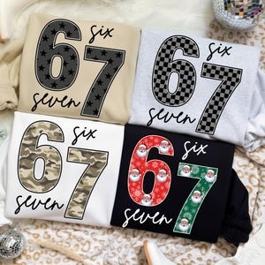 May include: Four sweatshirts with the numbers "67" and the word "six" and "seven" printed on them. The numbers are in different patterns: black stars, checkered, camouflage, and Santa Claus faces. The sweatshirts are in beige, gray, white, and black.