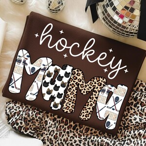 May include: A brown sweatshirt with the words "hockey mom" in white script. The letters "MOM" are filled with hockey-themed and leopard print patterns. The sweatshirt is folded on a surface with a leopard print pattern.