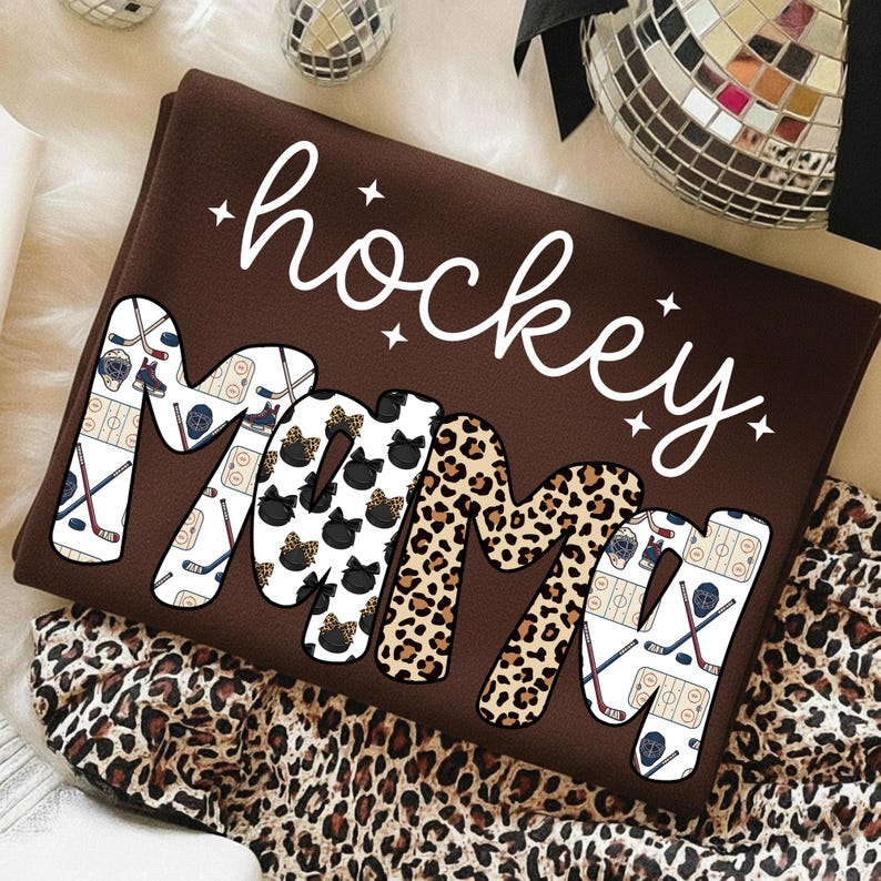 May include: A brown sweatshirt with the words "hockey mom" in white script. The letters "MOM" are filled with hockey-themed and leopard print patterns. The sweatshirt is folded on a surface with a leopard print pattern.