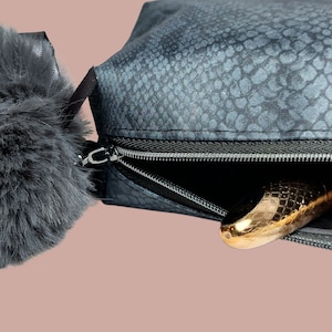 以下が含まれることがあります： スネークスキン模様のダークグレーのコスメティックバッグ。黒いジッパーとふわふわのグレーのポンポンが付いています。金色の物体が、開いたバッグの中から部分的に見えています。