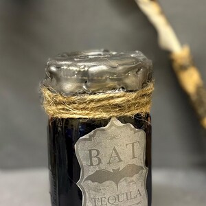 May include: A small glass jar filled with a dark liquid, sealed with a silver-colored top and twine. The jar has a label that reads "BAT TEQUILA" with a bat graphic. The label also states "Bottled in Transylvania Since 1888".