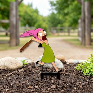 Könnte beinhalten: Ein flacher, zweidimensionaler Aufsteller einer Zeichentrickfigur mit schwarzen Haaren, die eine rosa Mütze mit Hasenohren und ein grünes Kleid trägt und einen braunen Baseballschläger schwingt. Die Figur hat einen weit aufgerissenen Mund und steht auf einem schwarzen Sockel.