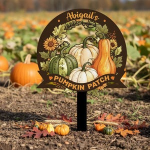 Peut inclure: Panneau décoratif de champ de citrouilles avec le texte "Abigail's Pumpkin Patch". Le panneau présente des illustrations de citrouilles, de tournesols et de feuillage d'automne. Le panneau est marron avec un piquet noir.