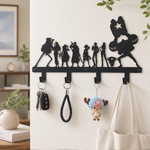Könnte beinhalten: Ein schwarzer Wand-Schlüsselhalter aus Metall mit Silhouetten von neun One Piece Anime-Figuren. Die Leiste hat vier Haken, an denen Schlüssel, ein geflochtenes Armband, ein kleiner Figuren-Schlüsselanhänger und eine beige Stofftasche hängen.