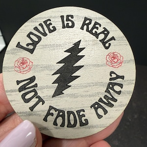 May include: Round wooden coaster with the text "LOVE IS REAL NOT FADE AWAY" in black lettering. A black lightning bolt design is in the center, with two red rose illustrations on either side. The coaster has a light wood grain background.