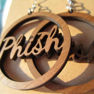 May include: Wooden hoop earrings with the word "Phish" cut out in a cursive font. The earrings are a light brown color and hang from silver-colored hooks. The earrings are displayed on a light brown surface.