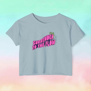 May include: Light blue t-shirt with the phrase "SURRENDER TO THE FLOW" in pink and a green frog graphic. The shirt has short sleeves and a crew neck. The design is centered on the front of the shirt.