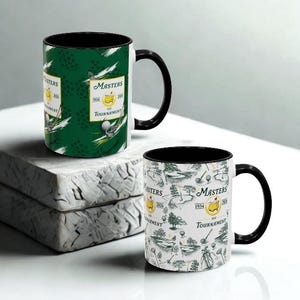 May include: Two ceramic coffee mugs with black handles and interiors. One mug features a green background with golf-themed graphics and the text "Masters 1934". The other mug has a white background with golf course illustrations and the same text.