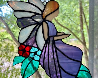 May include: A stained glass fairy with purple, white, and orange details. The fairy has large wings and is seated on a cluster of red, blue, and green leaves. The artwork is hanging from a window with a suction cup.