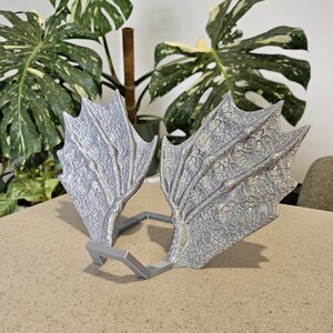 May include: Silver-coloured dragon wings with a textured, scale-like pattern. The wings are connected by a grey, angular frame, possibly for a costume or decoration. The wings are displayed on a neutral surface, with leafy green plants in the background.