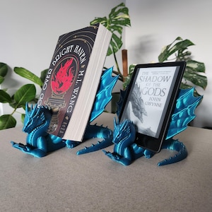 May include: Two teal dragon bookends supporting a book and a tablet. The book is titled "Bright Haven" with a red and gold cover. The tablet displays "The Shadow of the Gods". The dragon bookends feature detailed wings and scales.