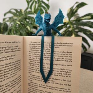 May include: A teal dragon bookmark sits on the pages of a book. The dragon has outstretched wings and is made of a textured material. The bookmark is a fantasy-themed item.