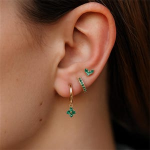 Könnte beinhalten: Eine Sammlung goldener Ohrringe mit smaragdgrünen Edelsteinen. Das Set umfasst einen kleinen Ohrstecker, einen Creolen mit einem hängenden vierblättrigen Kleeblatt-Design und einen kleinen Creolen mit grünen Steinen.