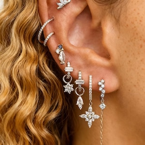 May include: An ear is shown with multiple pieces of jewellery. The ear has various earrings, including studs, hoops, and dangling styles, all with a sparkling, clear gemstone design. The jewellery is set against the backdrop of wavy, light brown hair.