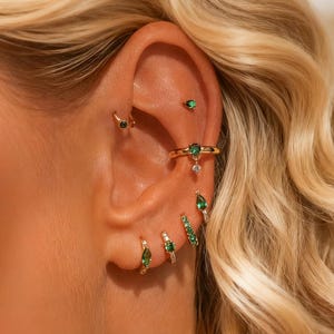 Pode incluir: Close-up de uma orelha adornada com vários brincos dourados com pedras preciosas verde esmeralda. Os brincos incluem pequenos brincos, argolas e um ear cuff decorativo, criando um visual em camadas e elegante. A orelha está contra um fundo de cabelo loiro.