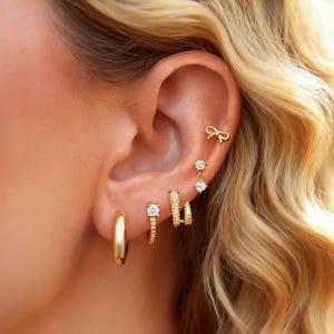 May include: Close-up of an ear with various gold earrings. The selection includes a small bow stud, a dangling pair with clear stones, a textured hoop, and a classic gold hoop. The earrings are set against a backdrop of wavy blonde hair.