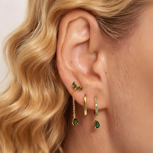 Può includere: Primo piano di un orecchio femminile adornato da tre orecchini d'oro. Uno presenta un design a foglia con pietre verdi, un altro è un semplice cerchio d'oro, e il terzo è un cerchio d'oro con pietre verdi e una goccia verde pendente.