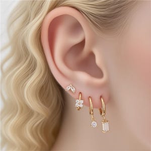 Könnte beinhalten: Nahaufnahme eines Ohrs, das mit mehreren goldenen Ohrringen geschmückt ist. Die Ohrringe umfassen einen kleinen Ohrstecker mit Blattdesign, einen blütenförmigen Creolen und zwei Creolen mit hängenden Edelsteinen. Die Edelsteine sind klar und funkelnd.