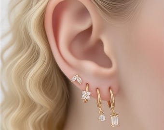 Könnte beinhalten: Nahaufnahme eines Ohrs, das mit mehreren goldenen Ohrringen geschmückt ist. Die Ohrringe umfassen einen kleinen Ohrstecker mit Blattdesign, einen blütenförmigen Creolen und zwei Creolen mit hängenden Edelsteinen. Die Edelsteine sind klar und funkelnd.