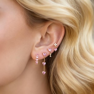 Könnte beinhalten: Nahaufnahme eines Ohrs, das mit mehreren Ohrringen geschmückt ist. Ein Ohrring hat drei rosa herzförmige Steine, ein anderer ein hängendes Design mit drei rosa Herzen und ein dritter ein kleines Blumenmuster. Die Ohrringe sind goldfarben.