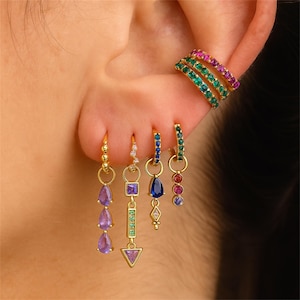 Op de afbeelding: Een verzameling gouden oorbellen en oormanchetten met kleurrijke edelstenen. De oorbellen hebben verschillende vormen, waaronder druppels, vierkanten en driehoeken, versierd met paarse, blauwe, groene en roze stenen. De oormanchetten zijn gestapeld met groene en roze edelstenen.