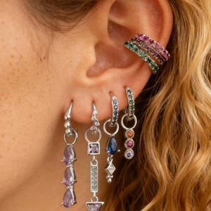 Pode incluir: Uma variedade de brincos e punhos de orelha. Os brincos apresentam vários designs, incluindo gotas, formas geométricas e pedras preciosas penduradas em tons de roxo, azul e rosa. Os punhos de orelha são empilhados, com fileiras de pedras preciosas coloridas.