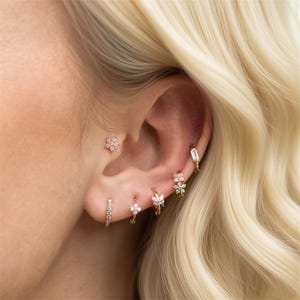 Puede incluir: Una oreja está adornada con una colección de pendientes de oro rosa. Incluyen pequeños aros con piedras transparentes, tachuelas de flores y una piedra rectangular en un aro. Los pendientes se colocan sobre un fondo de cabello rubio.