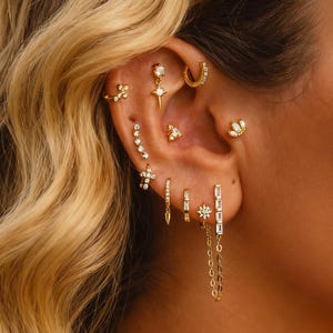 Op de afbeelding: Close-up van een oor versierd met meerdere gouden oorbellen. De oorbellen hebben verschillende ontwerpen, waaronder studs, hoepels en bungelende stijlen met heldere edelstenen. Het oor staat tegen een achtergrond van golvend blond haar.