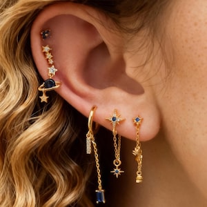 May include: A collection of gold-toned earrings featuring celestial designs. The earrings include star and planet motifs, with blue and clear gemstone accents. Some earrings dangle with chain details, showcasing a variety of shapes and sizes.
