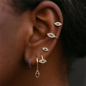 May include: A collection of gold-toned earrings, including hoops, studs, and a cuff, all featuring blue and clear gemstones. The earrings are arranged on an ear, showcasing various designs and styles. The jewelry is set against a dark background.