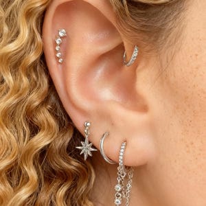 Pode incluir: Close-up de uma orelha adornada com vários brincos de prata. Um brinco de argola curvo, um brinco pendurado em forma de estrela e um brinco de corrente com três pedras preciosas penduradas são visíveis. Uma série de pequenas pedras preciosas ascendentes também são colocadas na orelha.