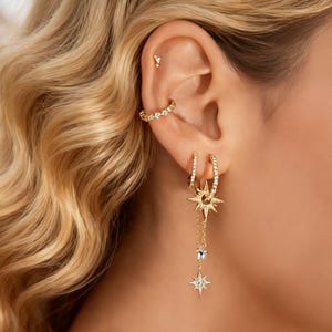 Puede incluir: Primer plano de una oreja adornada con pendientes dorados. Los pendientes incluyen un pequeño pendiente de botón, un delicado brazalete para la oreja y pendientes de aro con detalles de estrellas y cristales. La joyería se encuentra sobre un fondo de cabello rubio ondulado.