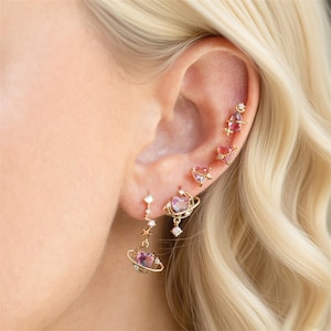 Puede incluir: Una colección de pendientes dorados, con diseños celestiales y de corazón. Los pendientes incluyen un pendiente colgante en forma de planeta con una piedra preciosa rosa, pendientes de corazón y un pendiente con acento de estrella. Los pendientes están engastados con piedras preciosas rosas y transparentes.